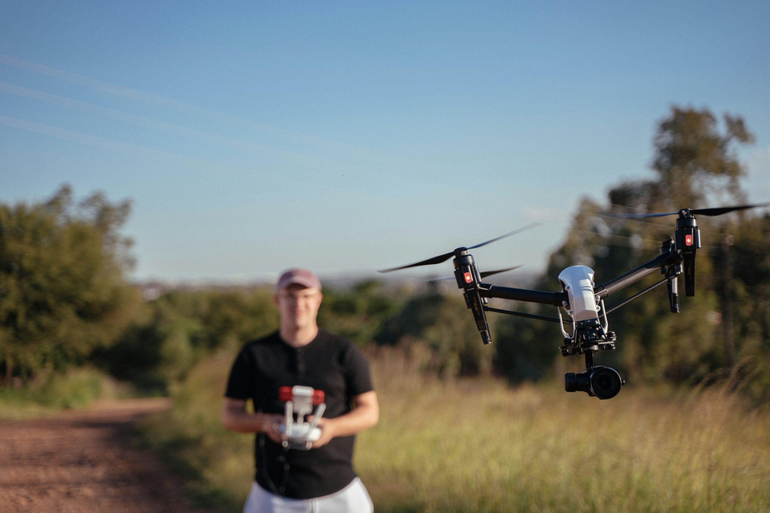 Drone with a mounted camera hovering in mid-air, controlled by a pilot standing in the background on a dirt path surrounded by greenery.